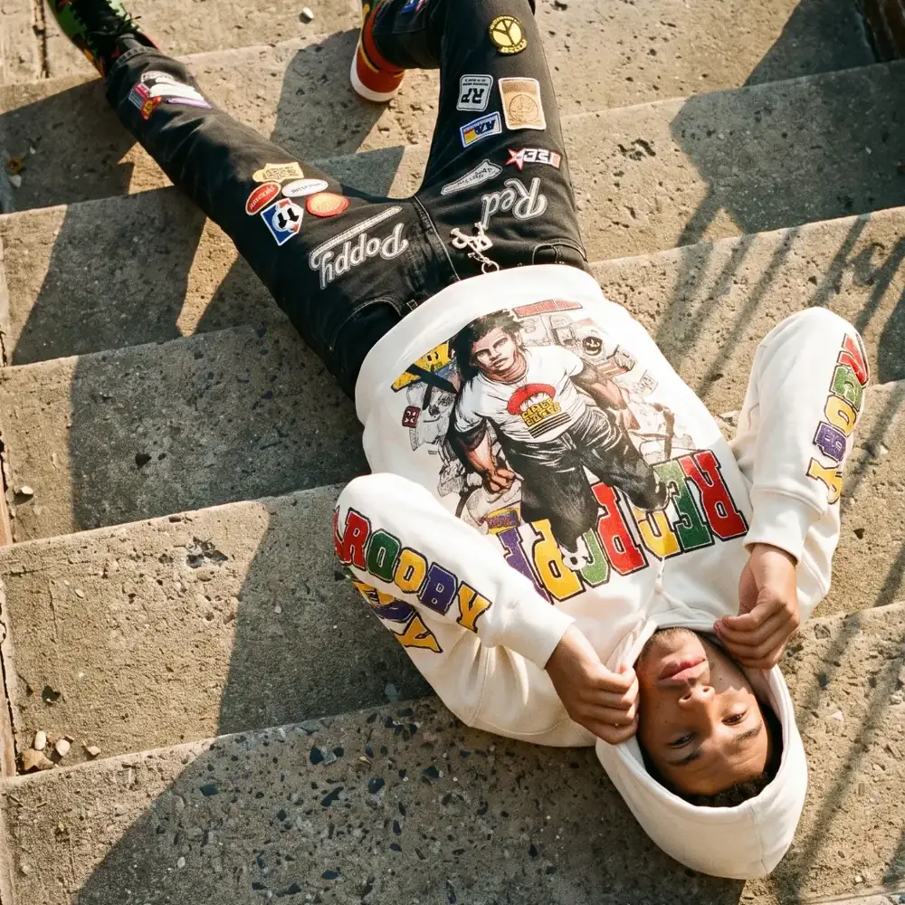 A model lying on concrete steps in a cream Red Poppy hoodie and black Red Poppy jeans covered in patches